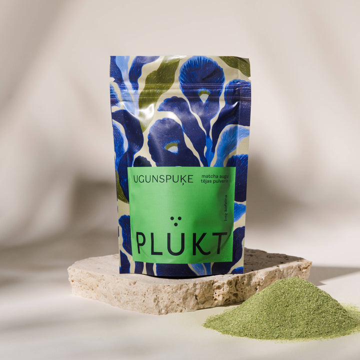 A stand-up pouch of Plukt Ugunspuke matcha green tea powder, featuring a blue and green floral pattern with a solid green label, displayed on a stone slab next to a small pile of the green powder.